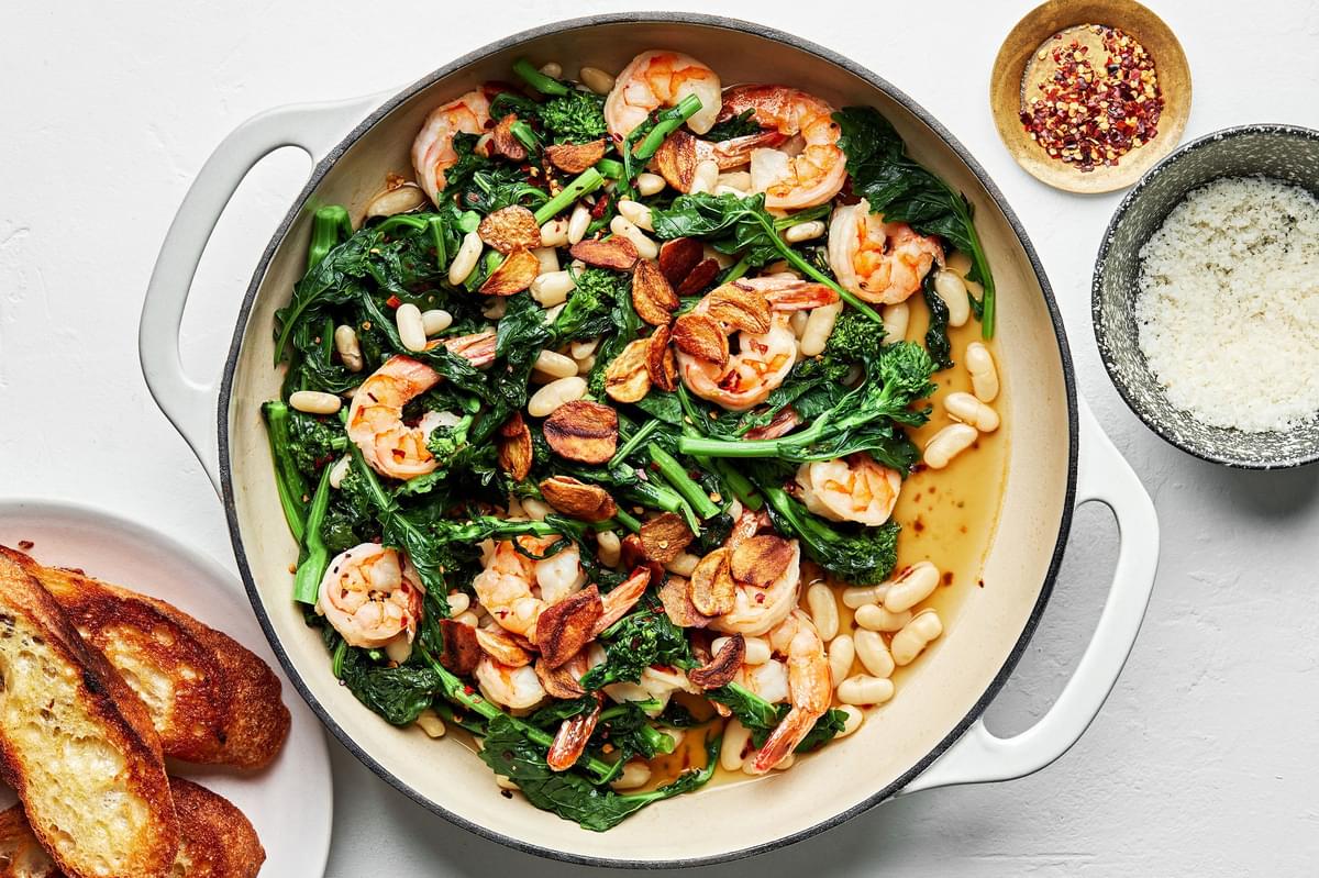 Spicy Broccoli Rabe with White Beans and Shrimp in a pan surrounded by red pepper flakes, parmesan & crusty bread for serving