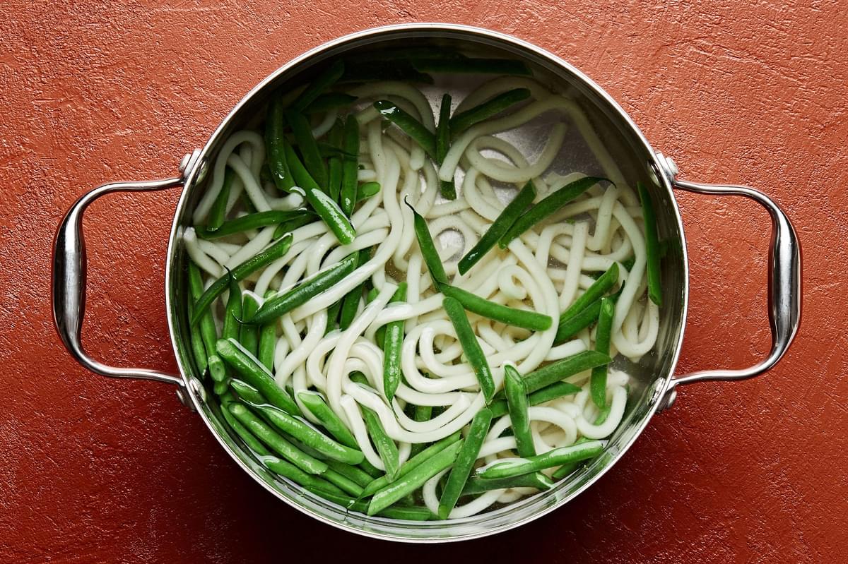 udon noodles and green beans being cooked in a pot of boiling water