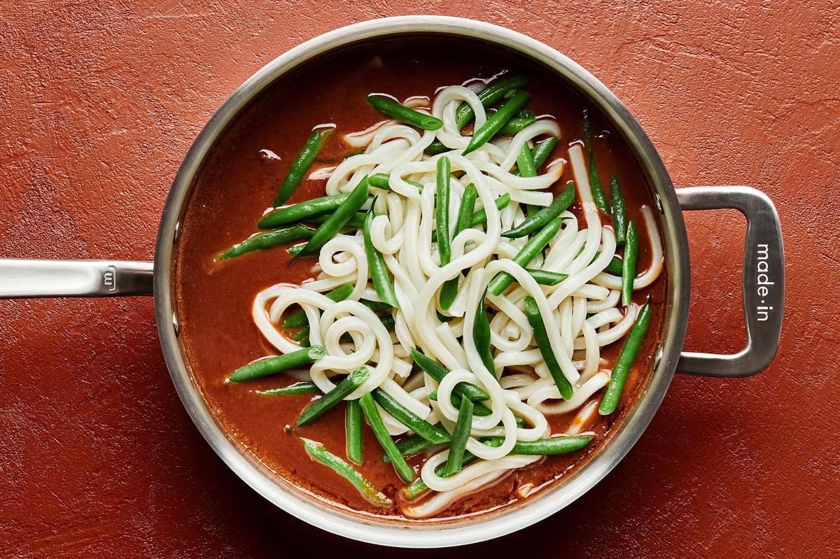 cooked udon noodles and green beans being tossed in a pan with spicy peanut sauce
