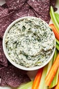 a bowl of homemade spinach dip surrounded by blue corn tortilla chips, carrot sticks and celery sticks for dipping
