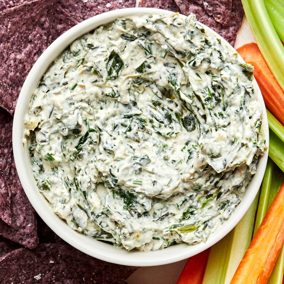 a bowl of homemade spinach dip surrounded by blue corn tortilla chips, carrot sticks and celery sticks for dipping