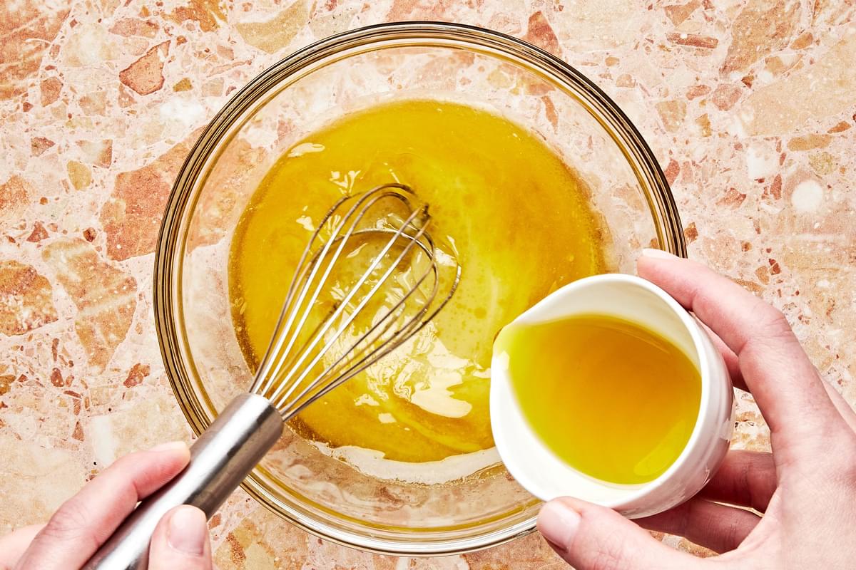 olive oil being whisked into a bowl with lemon juice, mustard, sugar, and salt