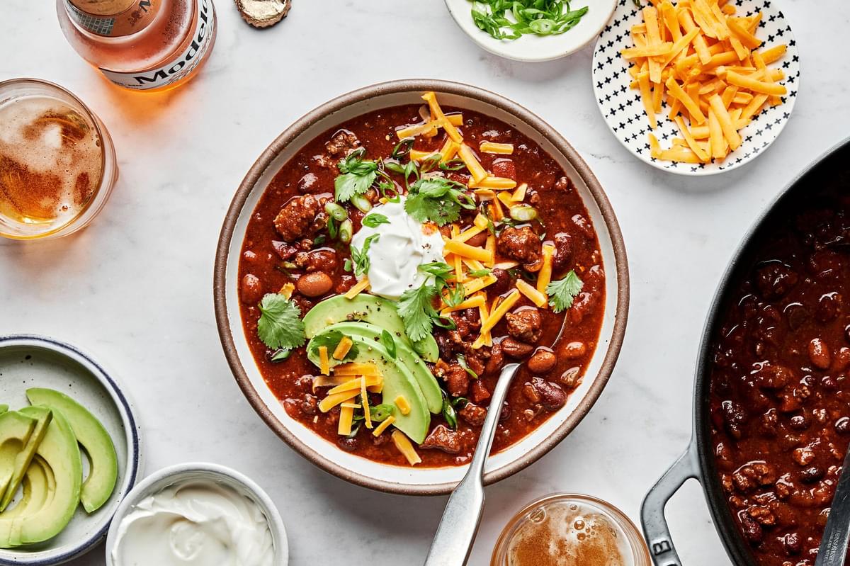 a bowl of homemade chili made with beef, pork, kidney, black and pinto beans topped with cheese, sour cream and avocado