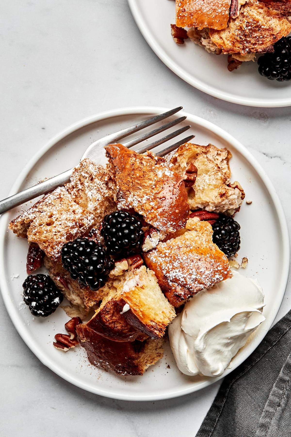 baked french toast topped with maple syrup, powdered sugar, whipped cream and berries on a plate with a fork