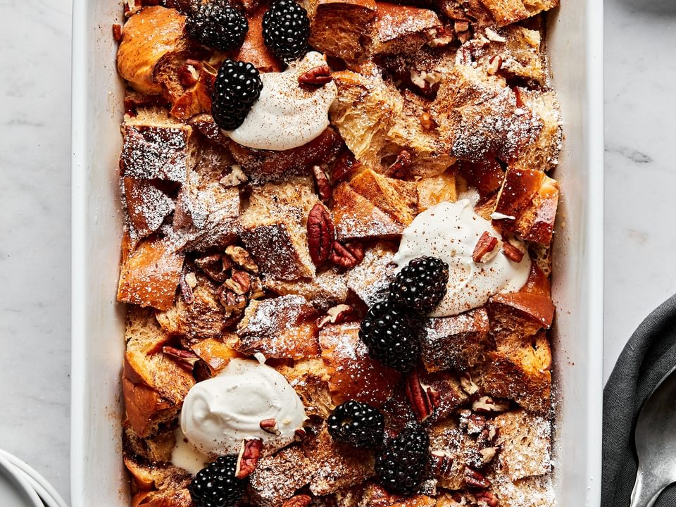 homemade baked french toast made with challah bread in a baking dish topped with dollops of whipped cream and blackberries