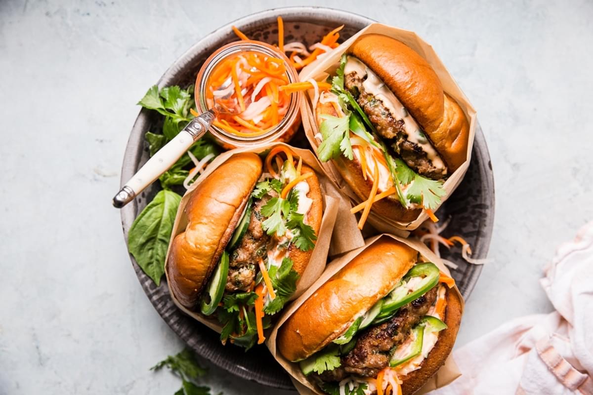 3 pork banh mi burgers on a plate with pickled veggies, cilantro and basil