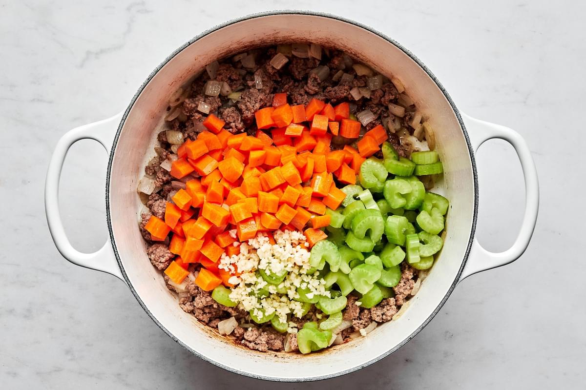 ground beef, chopped onion, carrots and celery and minced garlic being cooked in a large pot with olive oil.