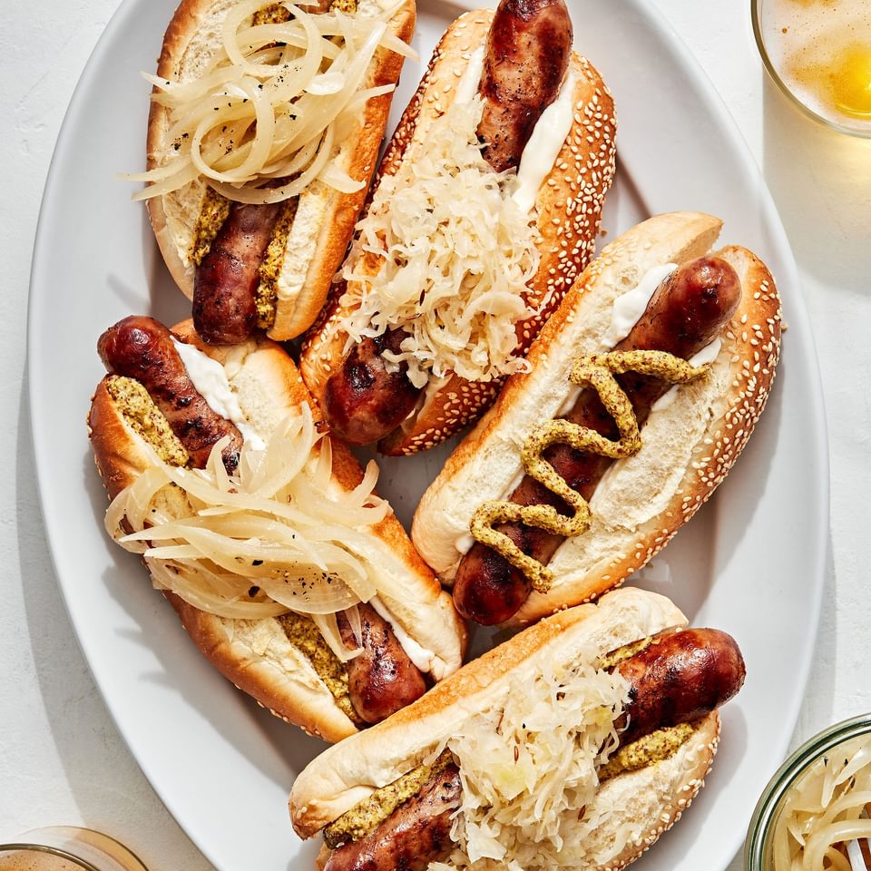 a platter of beer brats served on hoagie rolls topped with sauerkraut, onions, mustard and mayo
