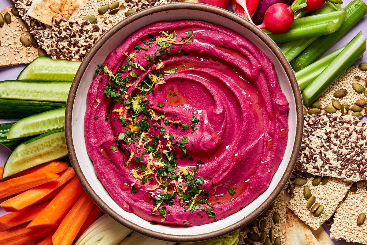 homemade beet hummus in a serving bowl surrounded by crackers and raw vegetables for dipping