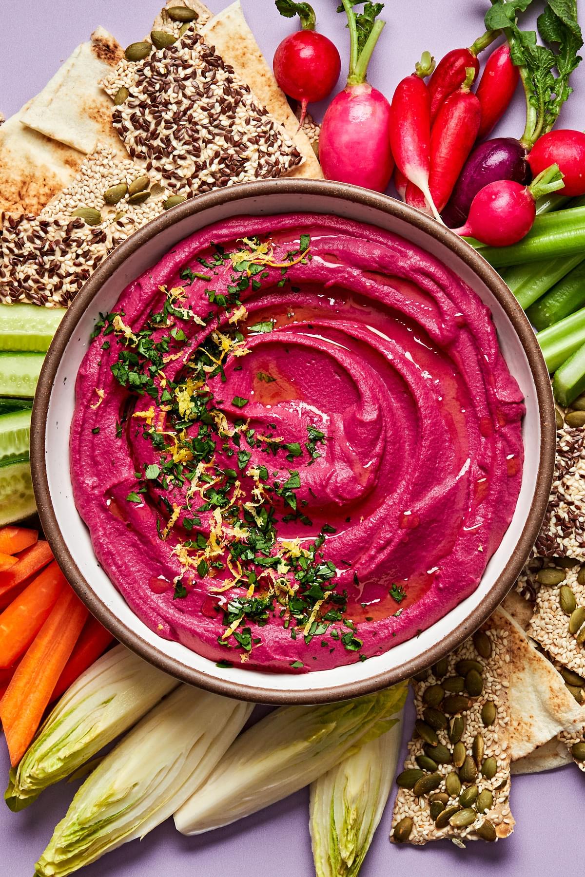 homemade beet hummus in a serving bowl surrounded by crackers and raw vegetables for dipping