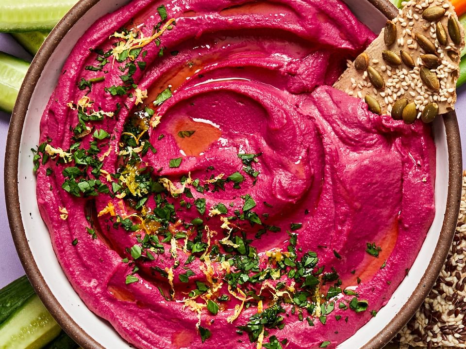 homemade beet hummus in a serving bowl surrounded by crackers, celery and carrots for dipping