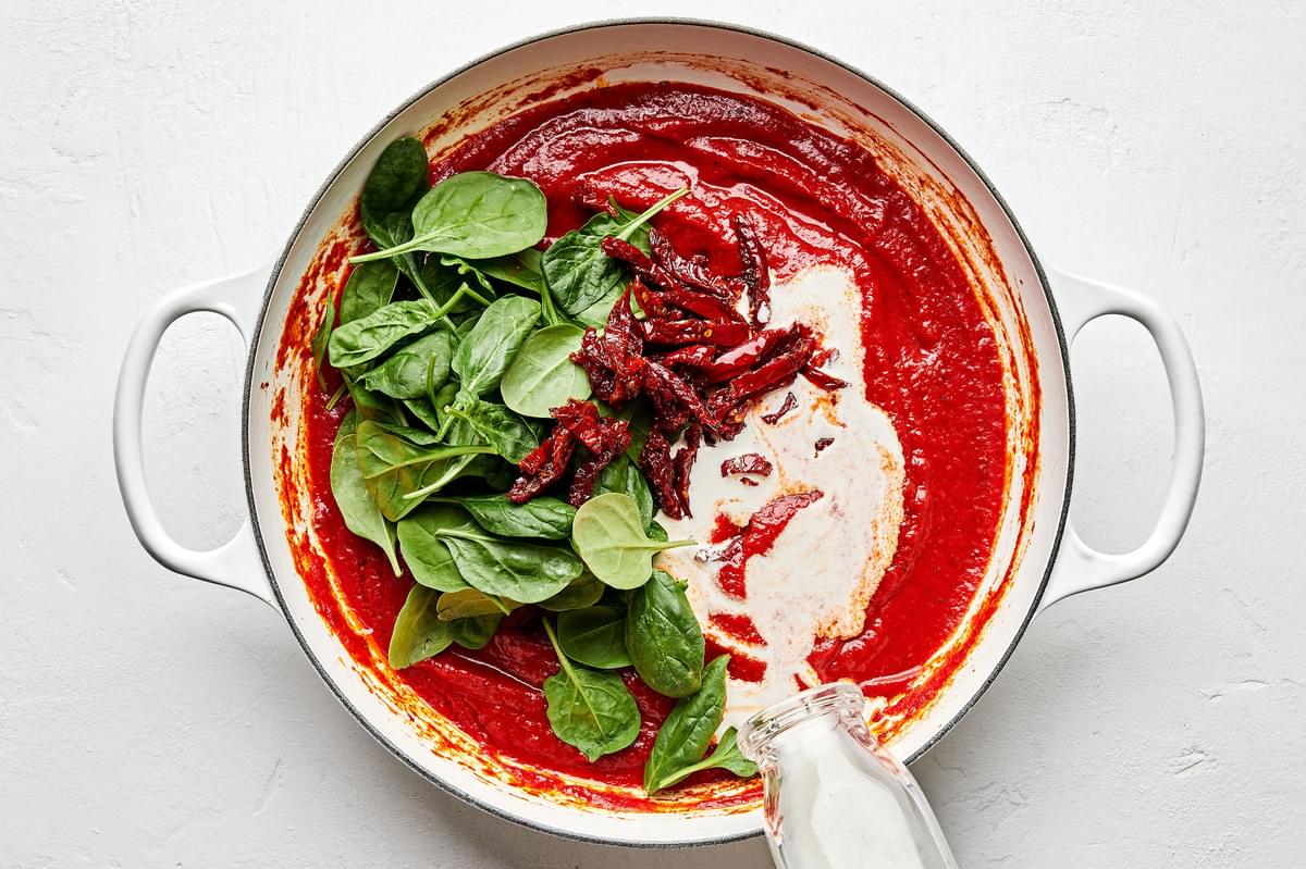 cream, sun-dried tomatoes & heavy cream being added to a pot with garlic, olive oil, tomatoes, spices & basil leaves