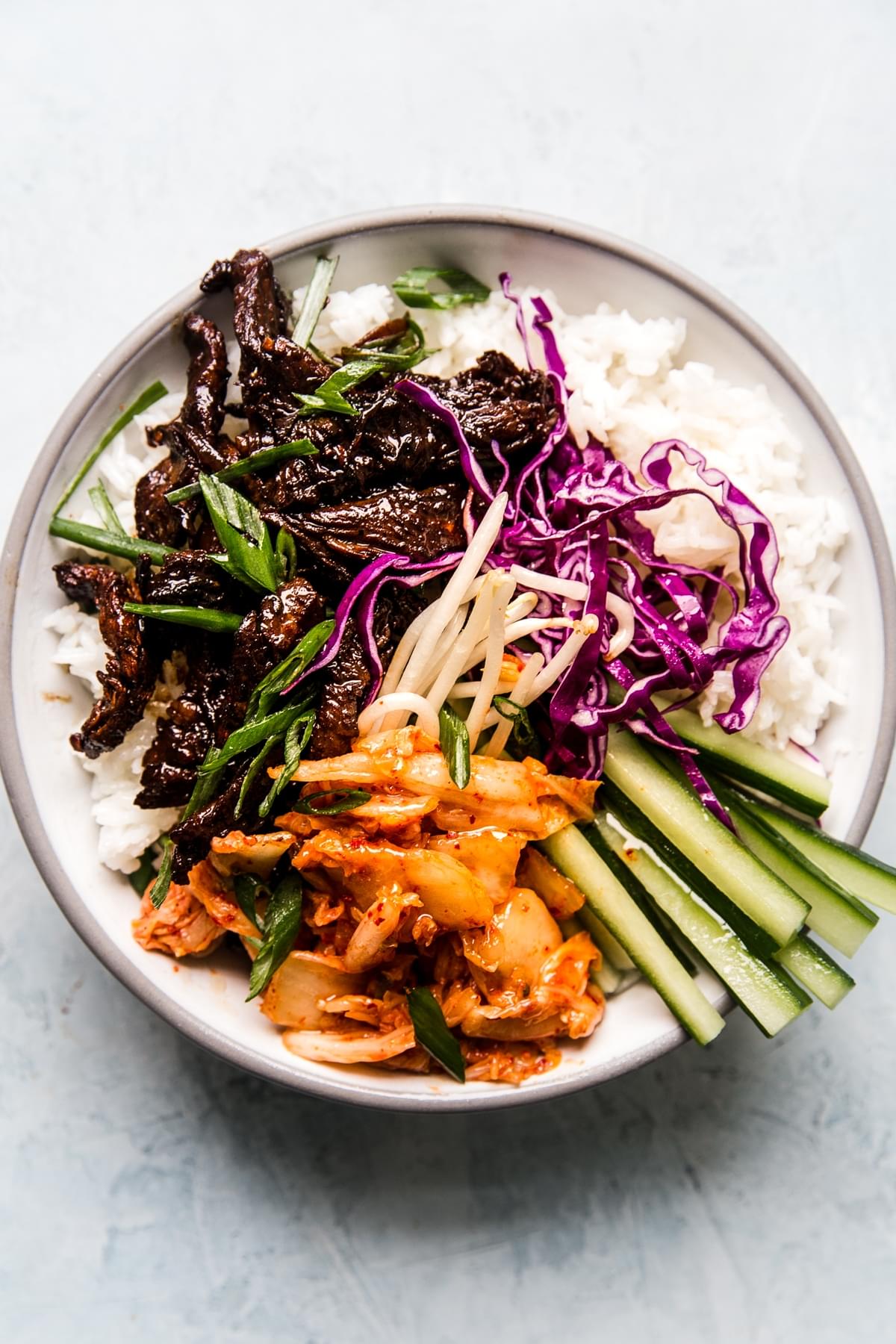 a bowl of white rice topped with beef bulgogi, kimchi, bean sprouts, red cabbage and cucumbers