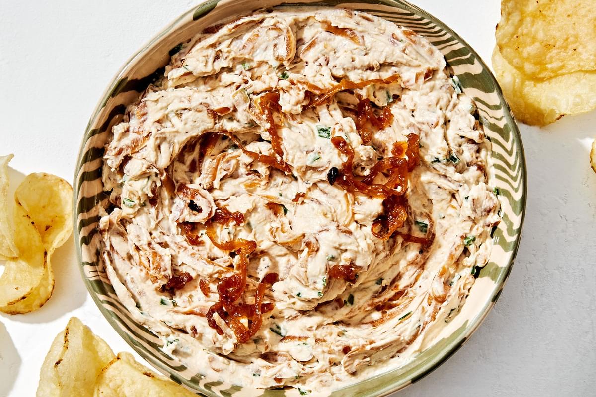 a bowl of caramelized onion dip made with cream cheese, sour cream, spices and chives surrounded by potato chips