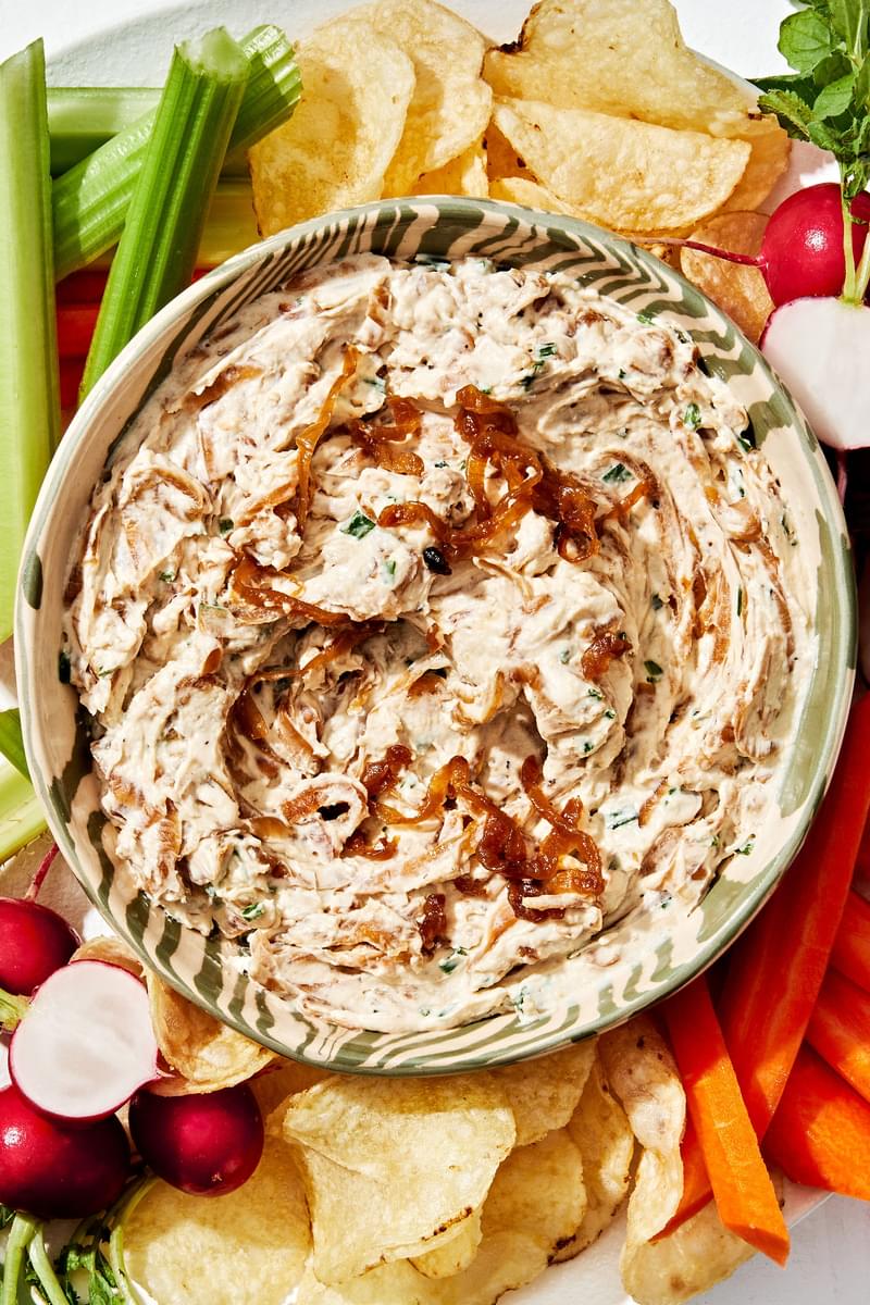 a bowl of homemade caramelized onion dip surrounded by potato chips and raw veggies for dipping