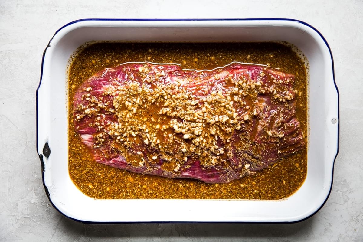flank steak being marinaded in a baking dish with garlic, orange juice, lime and spices for carne asada.