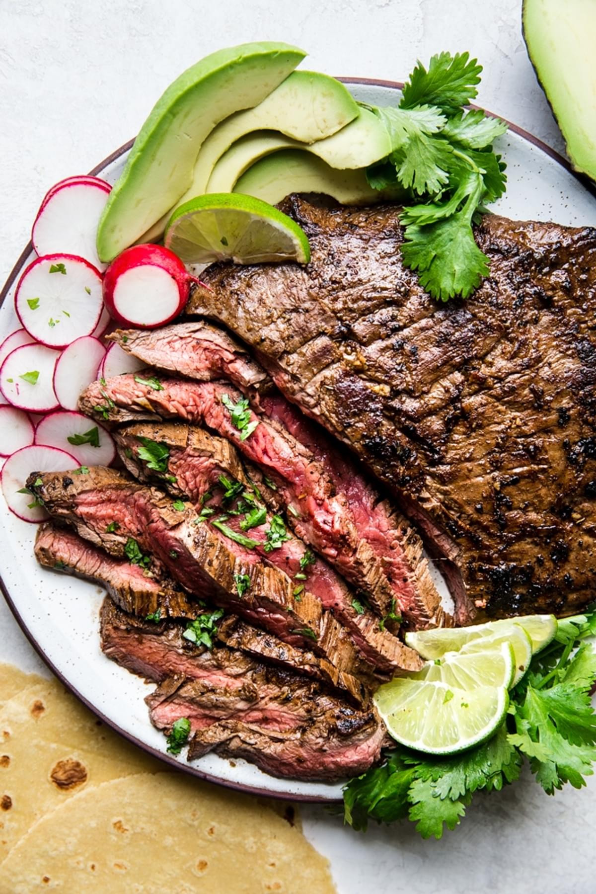 grilled flank steak for Carne asada on a plate with avocado, lime, radish, cilantro and tortillas