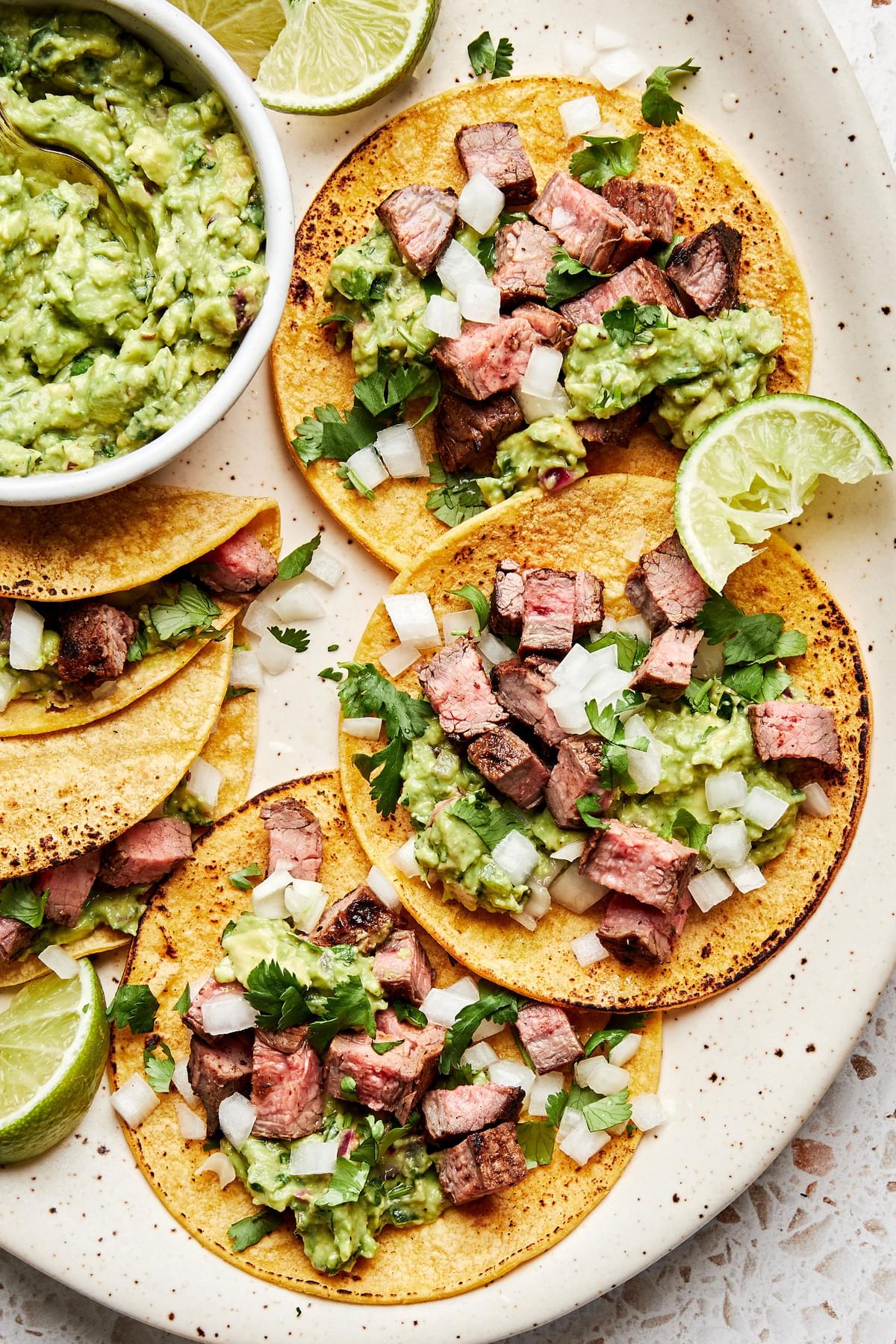 a plate of carne asada tacos topped with cilantro, onion, and guacamole served with a lime wedge