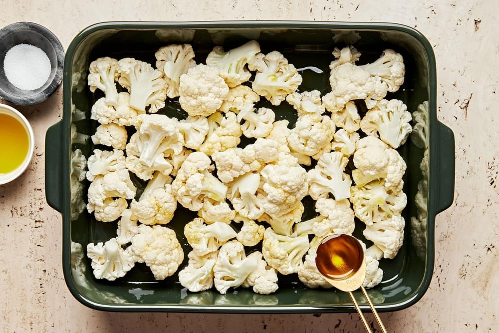 raw cauliflower florets being tossed with olive oil and salt in a baking dish