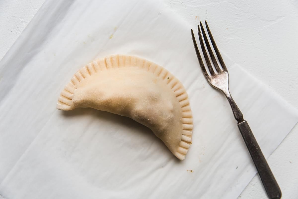 raw chicken empanada folded and pressed together with a fork on parchment paper