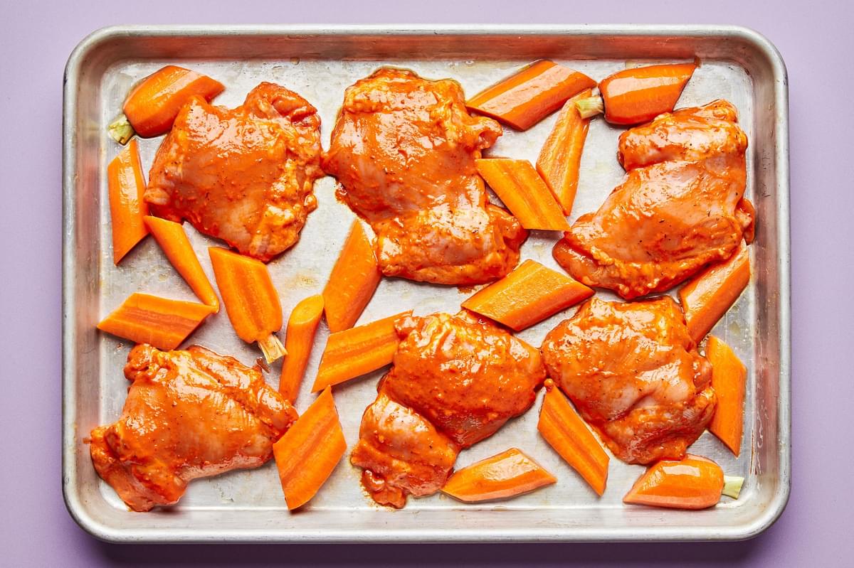 chicken thighs tossed in red pepper sauce and sliced carrots on a baking sheet ready to be roasted in the oven