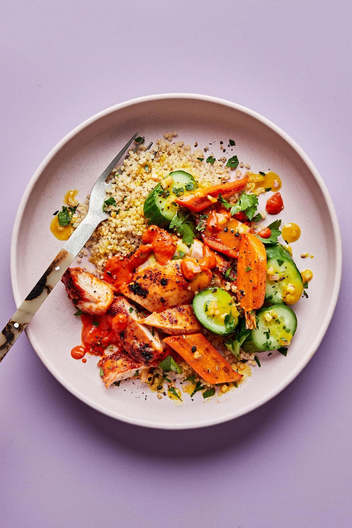a bowl of quinoa topped with chicken with red pepper sauce, roasted carrots, cucumbers and drizzled with turmeric vinaigrette