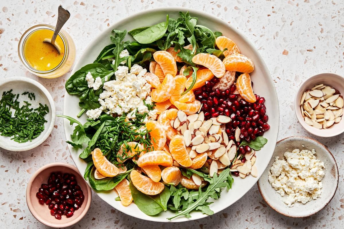 Christmas Salad being assembled with spinach, arugula, feta, clementines, pomegranates, almonds and chives