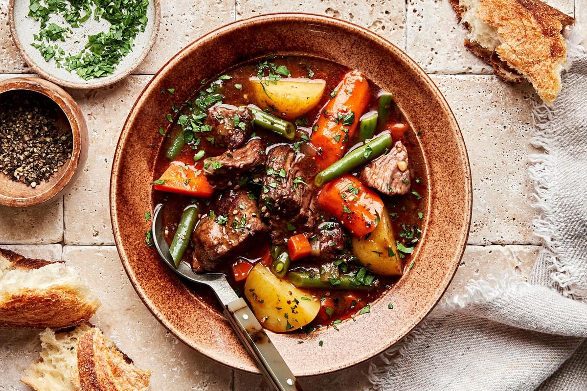 classic beef stew with green beans, potatoes and carrots sprinkled with parsley in a bowl with a spoon served with bread