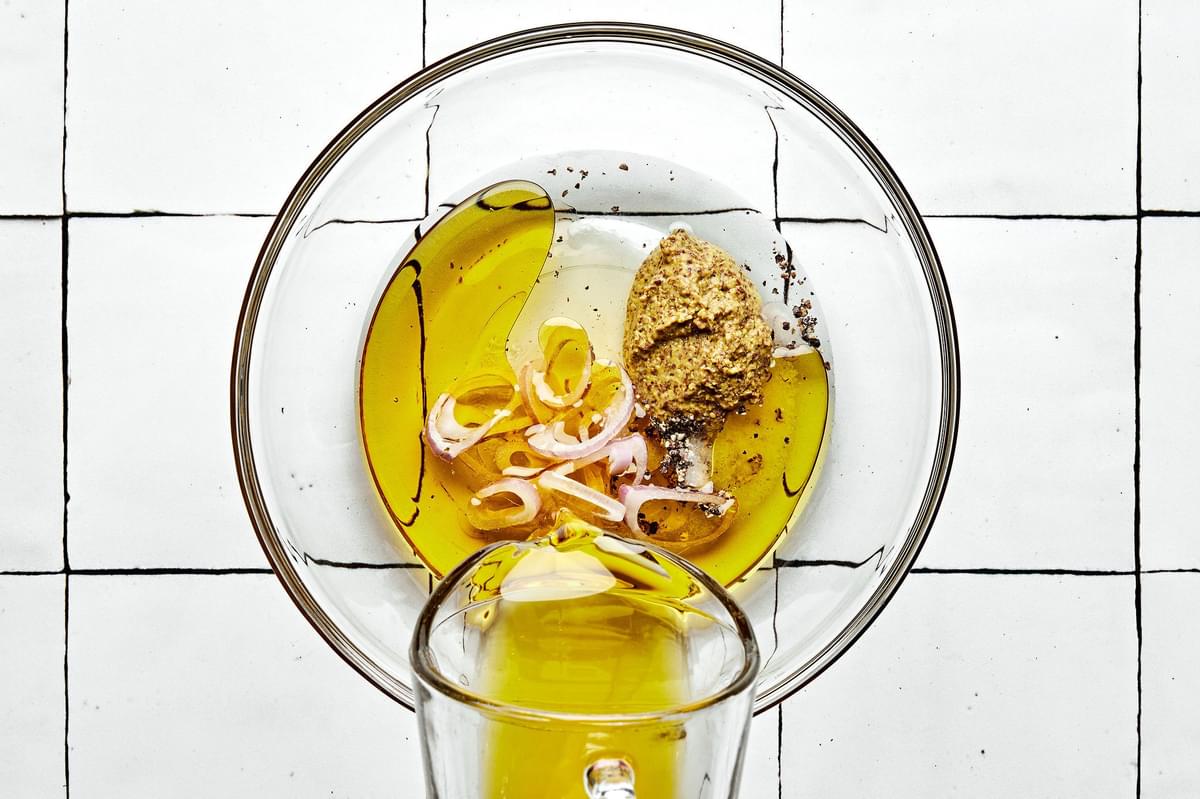 olive oil being poured into a bowl with sliced shallots, stone-ground mustard, honey, white wine vinegar, salt and pepper