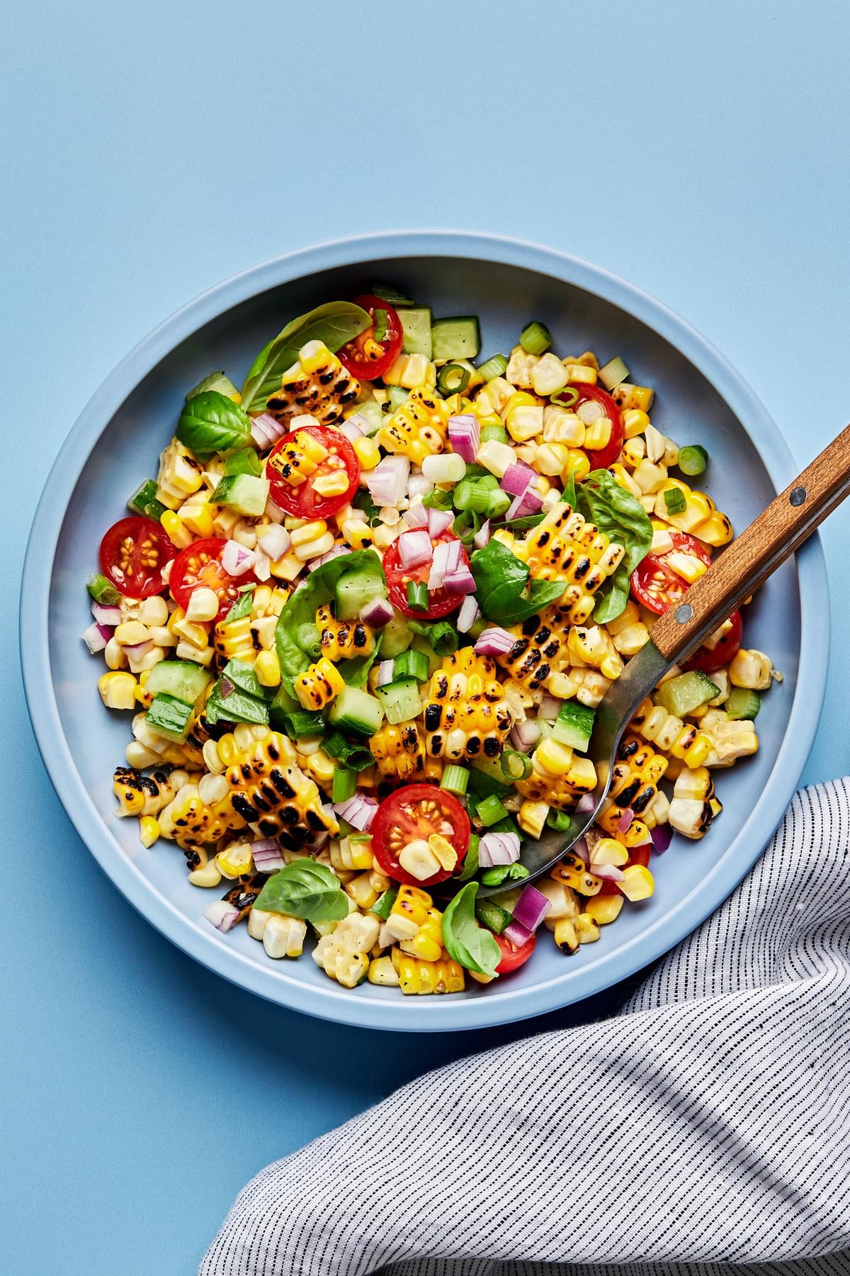 corn salad in a serving bowl with a spoon made with tomatoes, cucumber, red onion, green onions, basil and lime juice