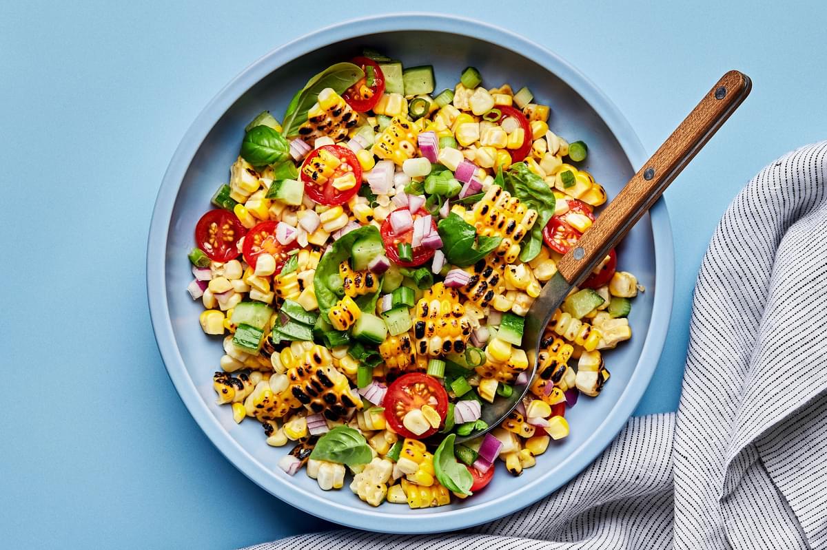 corn salad made with tomatoes, cucumber, red onion, green onions, basil and lime juice in a serving bowl with a spoon