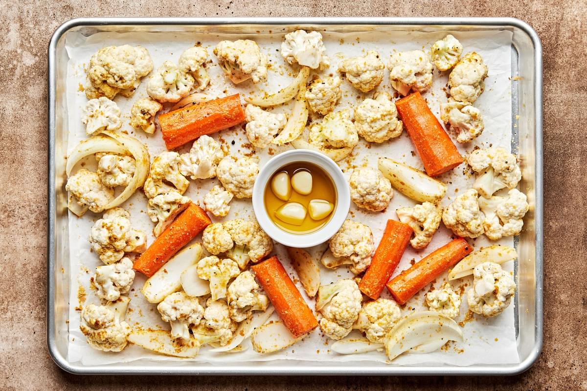 cauliflower, carrots & onion tossed with olive oil, nutritional yeast,  salt & pepper and a dish of garlic on a baking sheet