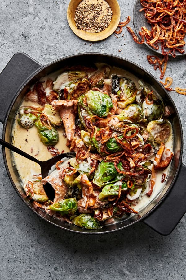 creamy brussels sprouts with wild mushrooms in a skillet with a serving spoon next to a bowl of fried shallots for topping