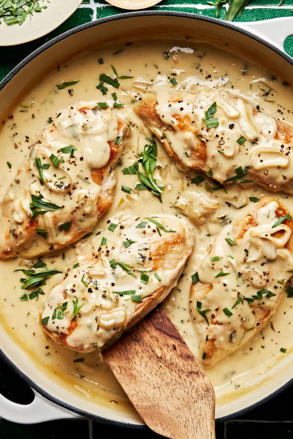 creamy chicken in a large skillet topped with tarragon and black pepper being scooped out with a wooden spoon.
