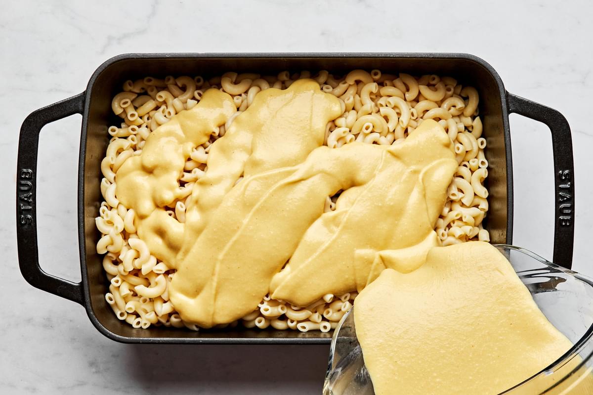 dairy free cheese sauce being poured over cooked macaroni noodles in a 9x13 pan