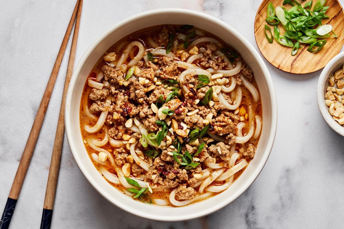 a bowl of homemade dan dan noodles sprinkled with green onions and crushed peanuts next to chopsticks