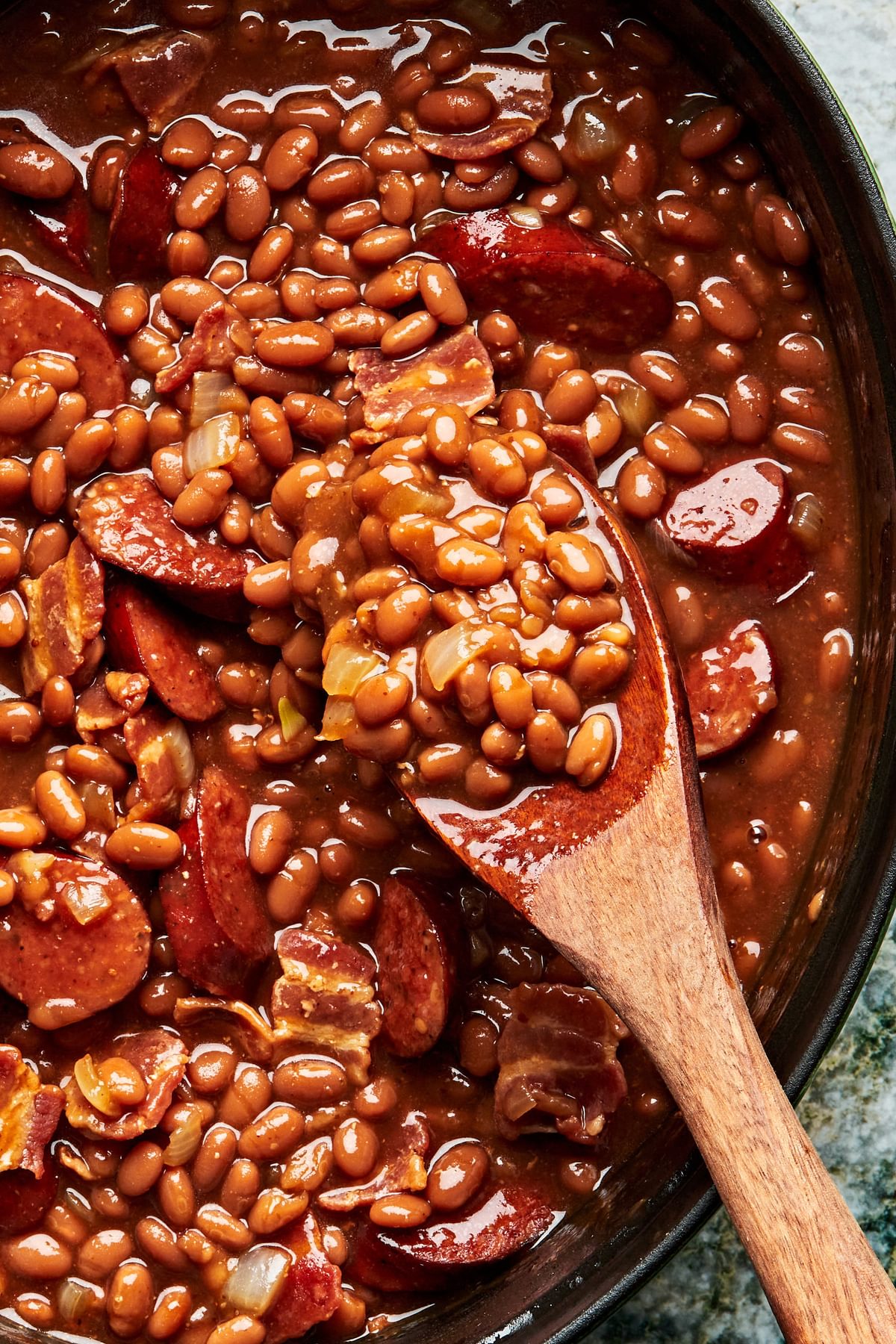 A large pot of baked beans with sausage, bacon, brown sugar, ketchup and mustard being stirred with a wooden spoon