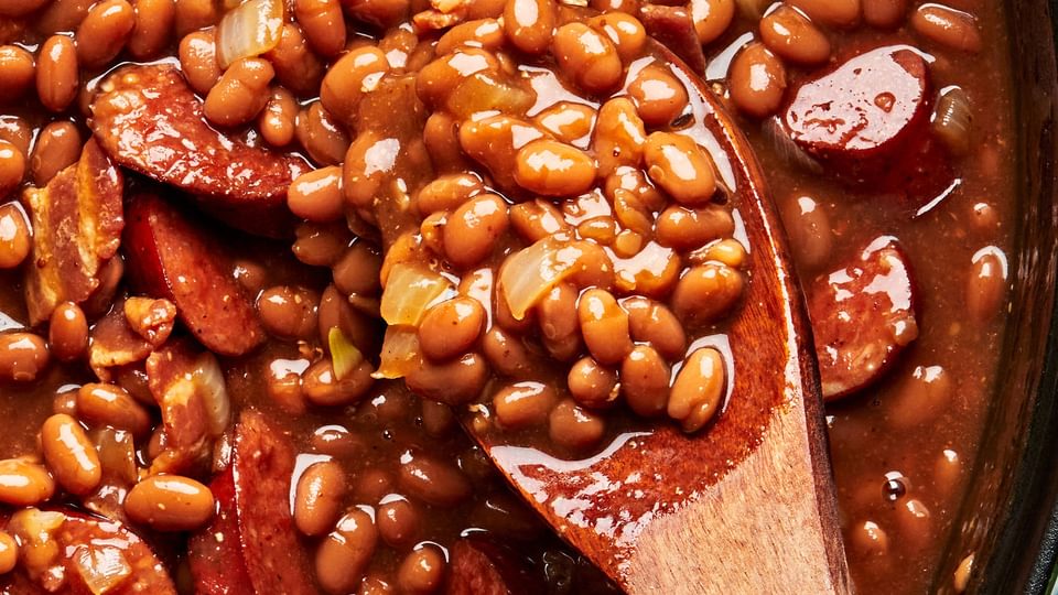 A large pot of baked beans with sausage, bacon, brown sugar, ketchup and mustard being stirred with a wooden spoon