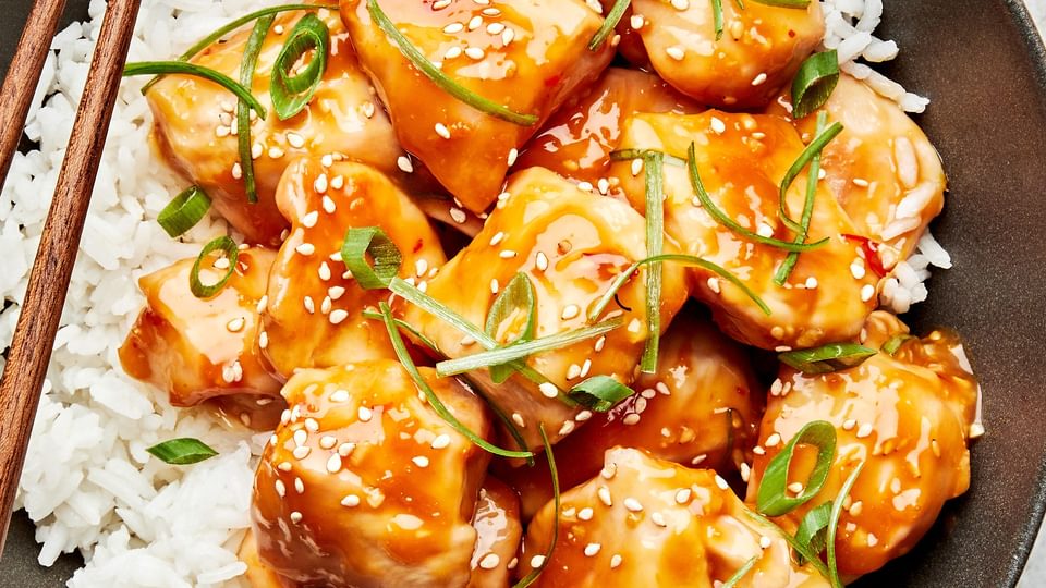 a bowl of orange chicken served on top of white rice and topped with sesame seeds and green onions