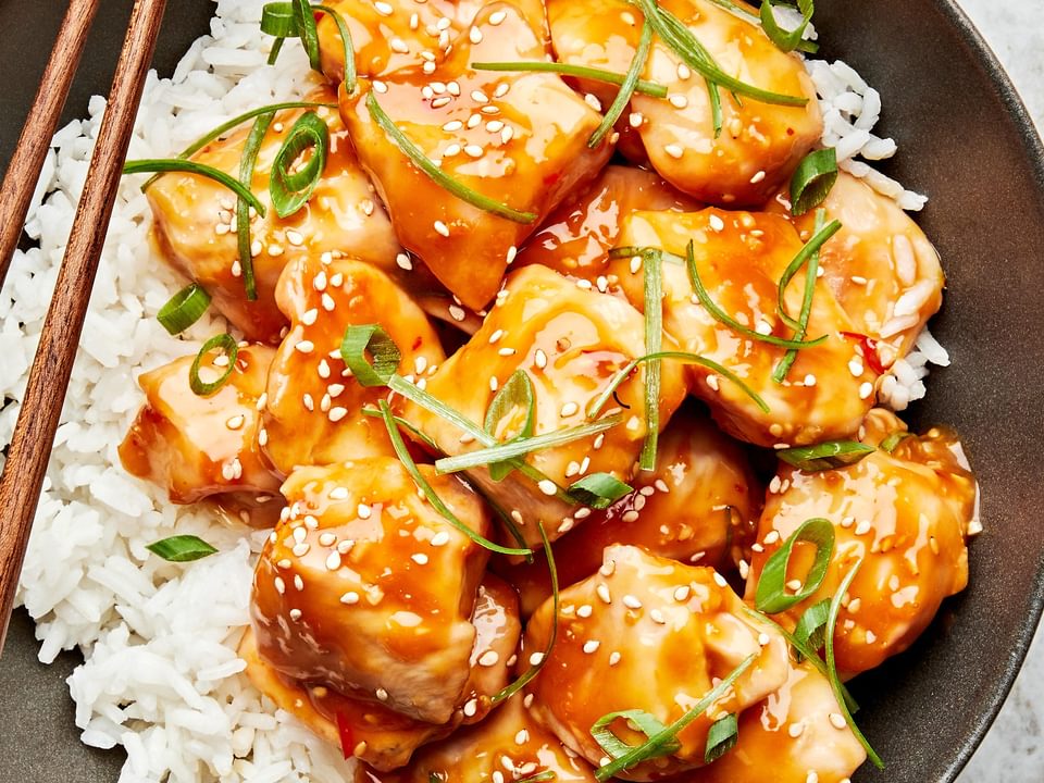 a bowl of orange chicken served on top of white rice and topped with sesame seeds and green onions