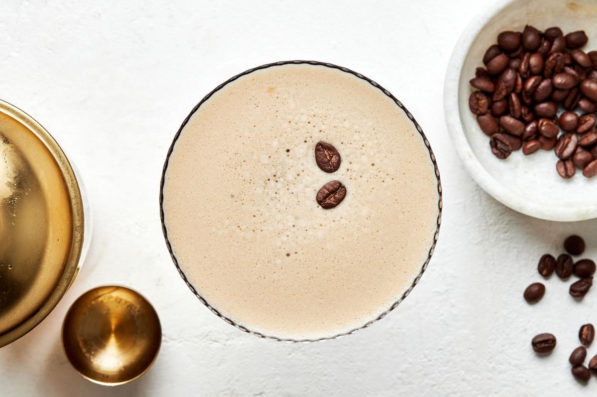 an espresso martini garnished with 2 coffee beans next to a bowl of additional coffee beans