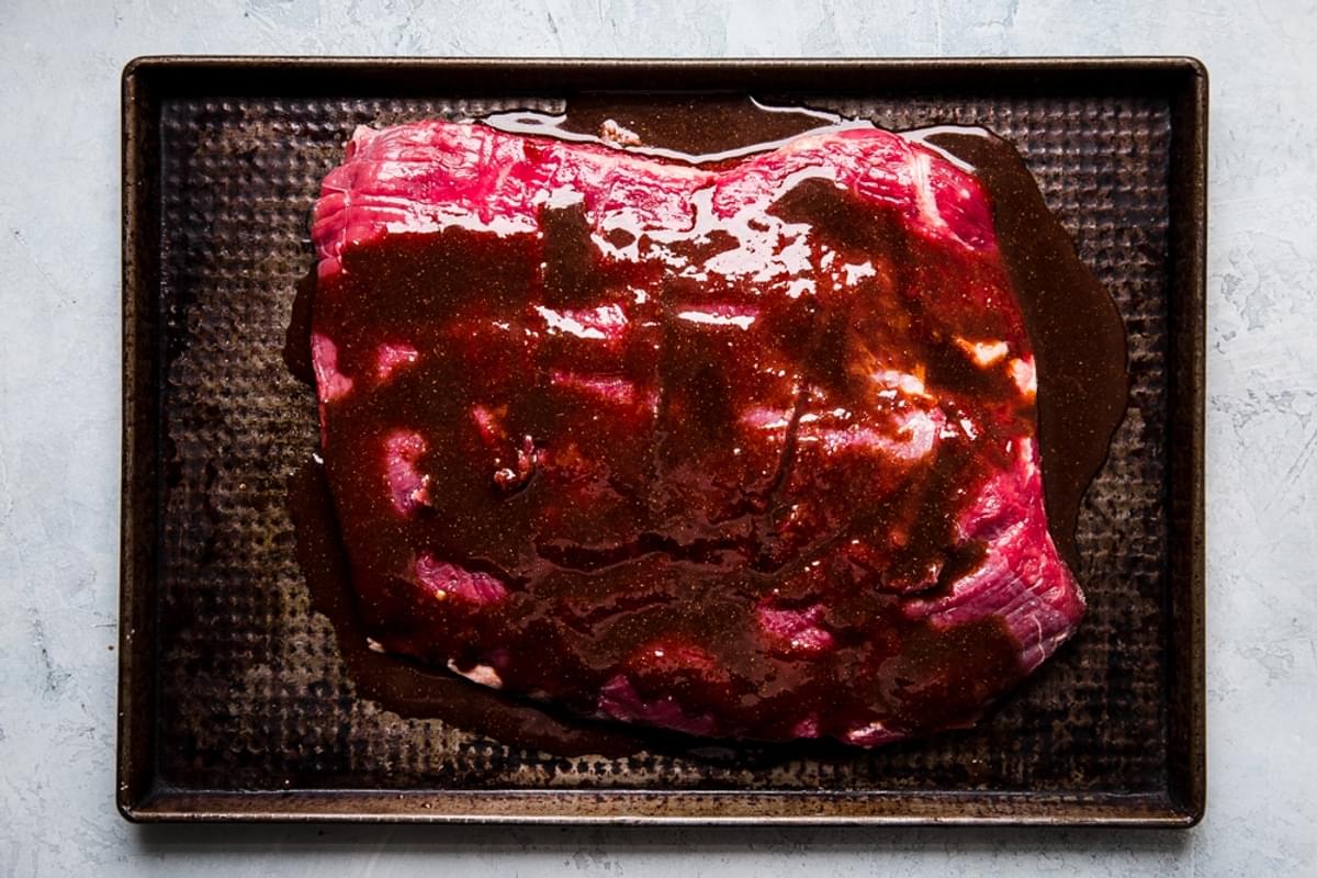 raw flank steak marinating on a rimmed baking sheet