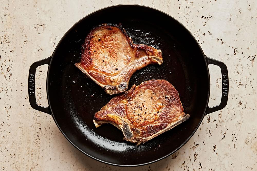 2 bone-in pork chops seasoned with salt and pepper being cooked in a skillet with vegetable oil