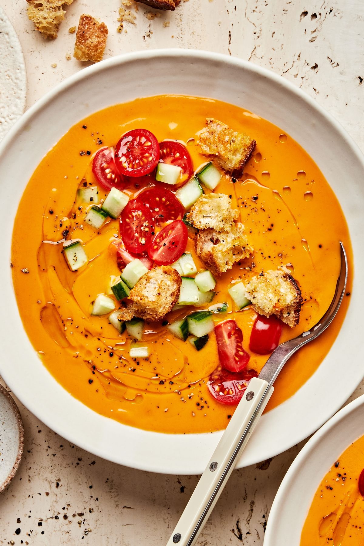 a bowl of gazpacho soup topped with a drizzle of olive oil, halved cherry tomatoes, diced cucumbers, croutons and pepper