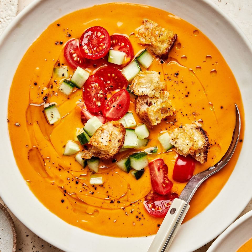 a bowl of gazpacho soup topped with a drizzle of olive oil, halved cherry tomatoes, diced cucumbers, croutons and pepper