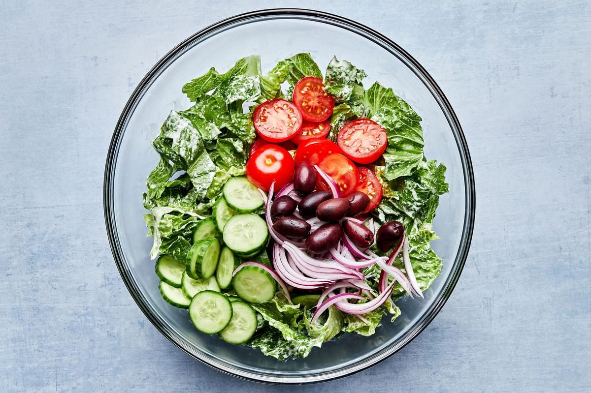 romaine, cherry tomatoes, persian cucumbers, kalamata olives and red onion being tossed in a glass bowl.