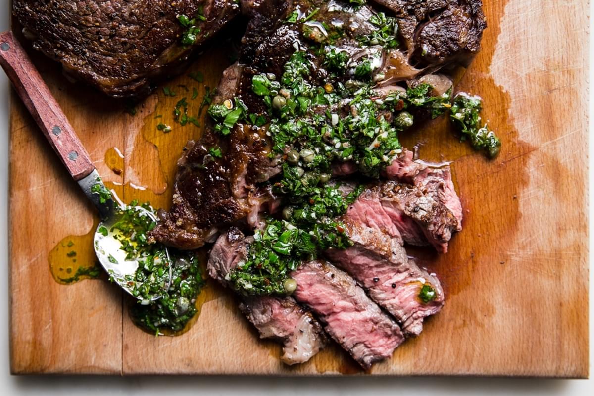 Grilled ribeye steak sliced on a cutting board with Italian salsa verde drizzled on top of it and on a spoon