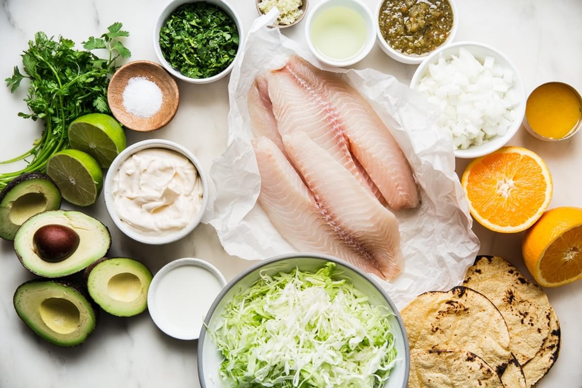 ingredients laid out for tilapia tacos, fish, tortillas, orange, lime, cilantro, mayonnaise, onions, avocado
