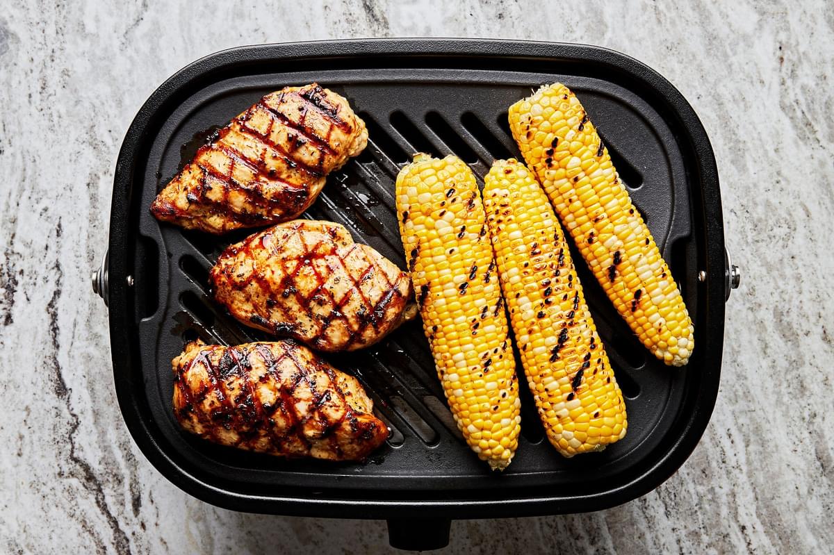 3 marinated chicken breasts and 3 corn on the cobs with char marks being cooked on a grill