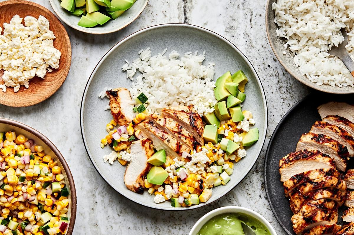 Grilled Chicken Bowl with corn and zucchini salad surrounded by avocado, feta and dressing in bowls